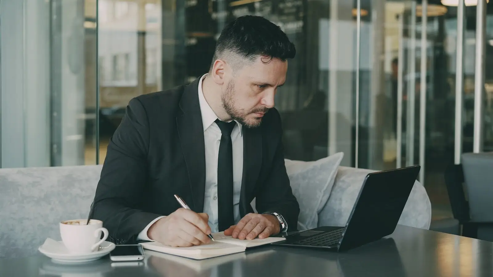 Man in suit writing in notebook at cafe table.